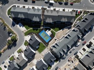 Aerial view of a residential area with row houses, a central tennis court, roads surrounding the buildings, and parked cars.