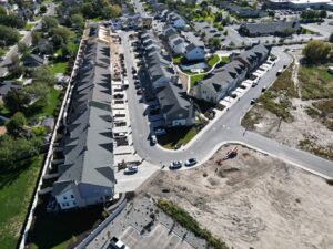 Aerial view of a suburban neighborhood with rows of houses, a parking area, and nearby undeveloped land.