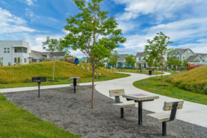 A park with a winding path, picnic benches, and BBQ grills showcases the perfect Daybreak setting against modern houses, reminiscent of the Contempo Collection, all under a clear blue sky.