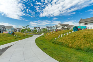 A winding path meanders through a grassy neighborhood park at daybreak, adorned with colorful sculptures and houses from the Contempo Collection under a vibrant blue sky.