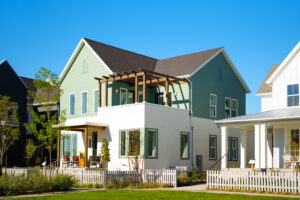 This modern two-story green house from the Contempo Collection features a white picket fence and a small balcony, all basking under the clear blue sky at daybreak.