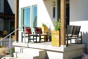 At daybreak, the sunny porch showcases wooden planters and black chairs with red cushions from the Contempo Collection, with house number 6669 standing proudly beside the door.