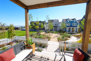 View from a porch with a bench, overlooking a pathway, gardens, and a row of colorful houses glowing under the clear blue sky at daybreak, as if touched by the vibrant hues of the Contempo Collection.