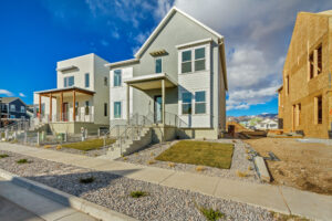 Modern residential street with two-story houses, one under construction, fronted by gravel landscaping and a sidewalk, under a blue sky.