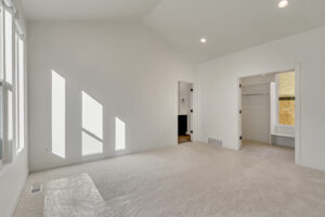 Empty room with beige carpet and white walls, featuring a closet and open door leading to another room. Sunlight streams in through three windows, casting shadows on the floor.