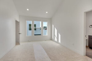 Empty room with light gray carpet, three large windows, and white walls. Room is well-lit with recessed ceiling lights. An open door leads to a bathroom.