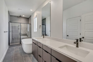 Modern bathroom with a freestanding tub, glass-enclosed shower, double sinks, and a large mirror. The room features grey tiles and minimalist decor.