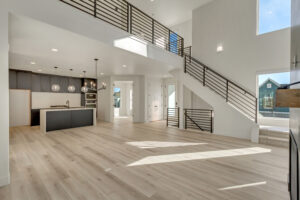 Spacious modern open-concept living area with a high ceiling, large windows, light wood flooring, a staircase with metal railings, and a kitchen with dark cabinets and pendant lights.