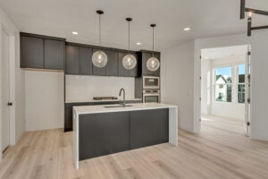 Modern kitchen with black cabinetry, white countertops, and three round pendant lights above an island. Light wood flooring and open doorway to a bright room with large windows.