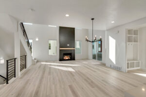 Spacious living room with light wood flooring, a modern chandelier, and a fireplace. Large windows allow natural light to fill the space.