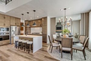 Modern kitchen and dining area featuring wood cabinetry, a large island with stools, stainless steel appliances, and a round dining table with chairs under a decorative chandelier.