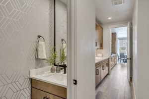 A small bathroom with geometric wallpaper, a white countertop, a rectangular mirror, and a black faucet and towel rack. It opens into a hallway leading to a kitchen area.