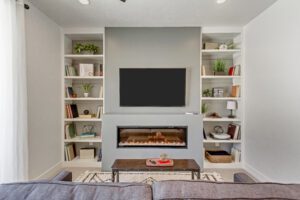 Modern living room with a wall-mounted TV above an electric fireplace, flanked by built-in bookshelves. The bookshelves feature books, decor, and small plants. A gray couch and coffee table are in the foreground.