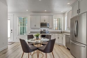Modern kitchen and dining area with stainless steel appliances, white cabinetry, a round glass dining table with four dark gray chairs, and a small potted plant centerpiece. Light wood flooring throughout.