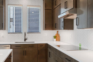 Modern kitchen in Cascade Village with dark cabinets, white countertops, a herringbone tile backsplash, and stainless steel appliances. This legacy design seamlessly combines contemporary aesthetics with timeless elegance.