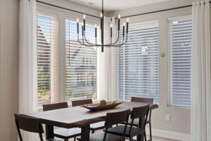 The dining area at Cascade Village features a wooden table, four chairs, and a modern chandelier, all embraced by the legacy of large windows with white blinds streaming natural light.