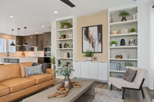 In the modern Cascade Village living room, a tan sofa sits beneath abstract art, complemented by bookshelves. A seamless view into the kitchen reveals elegant pendant lighting, blending contemporary design with legacy charm.