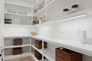 The modern pantry in Cascade Village features white shelves, wicker baskets, and jars, all beautifully illuminated by under-cabinet lighting on the sleek countertop.