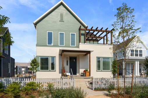 A modern two-story house in Cascade Village with a green upper facade, white lower facade, and a wooden pergola, surrounded by a white fence.