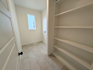 The empty pantry with white shelves and a tiled floor, reminiscent of a Cascade Village charm, features a window and a closed door.