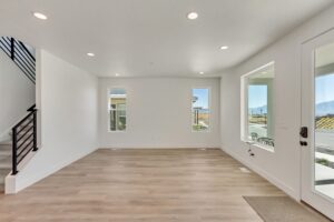 Empty room with light wood flooring, white walls, recessed lighting, and two windows. Black metal staircase railing on the left and a glass door on the right leading outside.