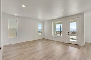 A spacious, empty room with light wooden flooring, white walls, recessed lighting, large windows, and a glass door leading outside.