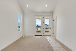 An empty room with beige carpet, white walls, and large glass doors leading to a balcony. A window on the left shows a view of mountains and houses. Plastic covers part of the floor.