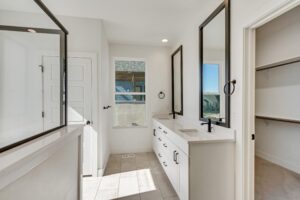 A bright, modern bathroom features a double vanity with two mirrors, black faucets, and white cabinets. It has a door, a window, and a tiled floor.