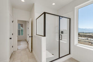 Bright bathroom with a glass shower enclosure, white tiles, and a large window overlooking a construction site. A partially open door leads to an adjacent room.