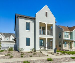 A row of modern, two-story houses with balconies, small yards, and a paved street under a clear blue sky.