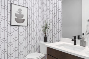 In the modern bathroom at Cascade Village, geometric wallpaper complements the white sink adorned with black fixtures, while curated wall art adds a touch of elegance. A plant on the toilet tank brings a hint of nature indoors.