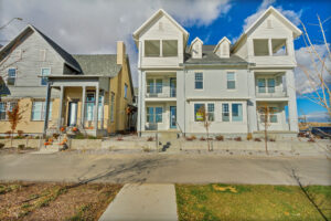 Two modern, three-story houses with gabled roofs and balconies, set against a clear sky. The foreground features a paved walkway and sparse landscaping.