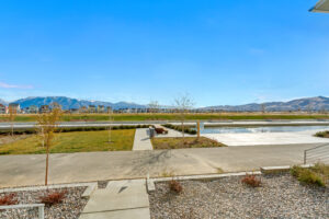 A paved walkway leads to a large open area with small trees and a water feature. Mountains are visible in the background under a clear blue sky.