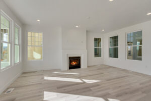 A bright, empty room with white walls, a fireplace, large windows, and light wood flooring.