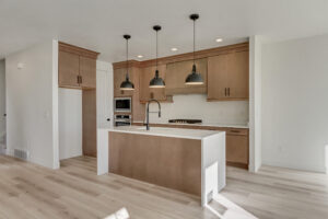 Modern kitchen with wooden cabinets, white countertops, and three black pendant lights above the island. Built-in oven and microwave are visible. Light-colored wooden floor.