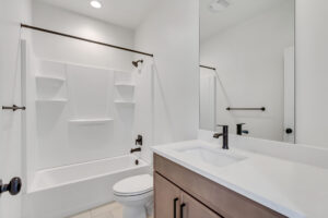 A clean, modern bathroom with a white bathtub, shower, toilet, and sink. Dark fixtures and a mirror above the sink enhance the minimalist design.