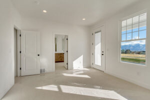 Bright, empty room with white walls and carpet, featuring two doors, a window with mountain views, and a doorway leading to a bathroom.