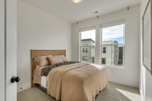 Cozy bedroom with a double bed, beige bedding, and two large windows showing an outdoor view of modern buildings. The space smartly incorporates basement automation for seamless control of lighting and ambiance.