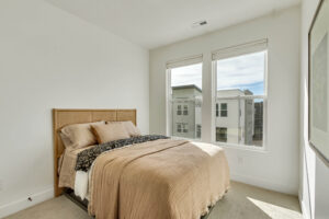 This minimalist bedroom features a neatly made bed with beige bedding and large windows offering an urban view, effortlessly blending style and function with ML+ enhancements for an optimized ambiance.