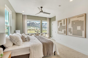 A modern bedroom with a large window offering mountain views, this space features neutral tones, abstract art, and a ceiling fan. Embrace tranquility in the style of ML+ with soft curtains that complete the serene ambiance upstairs, distinct from any basement vibes.