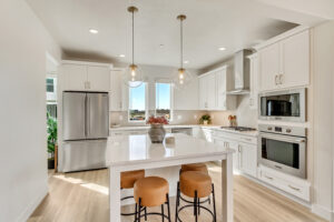 Modern kitchen with white cabinets, an island with stools, and pendant lights. Featuring stainless steel appliances and wood flooring, this space also boasts a convenient connection to the ML+ basement for seamless living and entertaining.