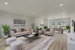 A modern living room with gray sofas, a glass coffee table, and indoor plants. Large windows provide natural light and a view of a green landscape.