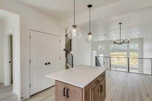 Modern kitchen with island, pendant lights, and a view of a bright living area overlooking Lake West through a large window with railing.