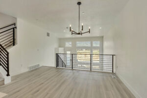 A modern living room with light wood flooring and a black railing staircase features a grand chandelier. Large windows provide a stunning view of Lake West, merging indoor elegance with the serene beauty of the water.