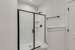 A minimalist bathroom with a glass-enclosed shower, white walls, and a black metal frame offers a serene escape reminiscent of the tranquility found at Lake West.