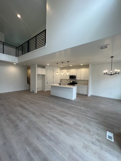 Spacious modern kitchen and living area with a high ceiling, light wood flooring, white cabinetry, an island with pendant lights, and a staircase with black railing.