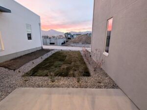 Small backyard with fresh sod and gravel border, flanked by two buildings. In the background, construction sites and a mountain range under a sunset sky.