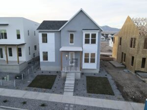 A newly constructed two-story house with a gray and white facade, concrete steps, and a small front lawn. Houses under construction are visible nearby.