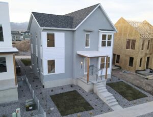 A two-story gray house with a porch, next to a similar house under construction. Gravel landscaping and some grass patches are visible.