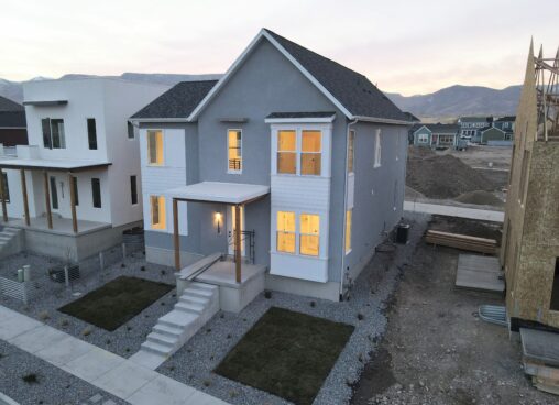 A two-story house with a grey and white exterior, illuminated windows, and a small landscaped yard in a residential construction area.